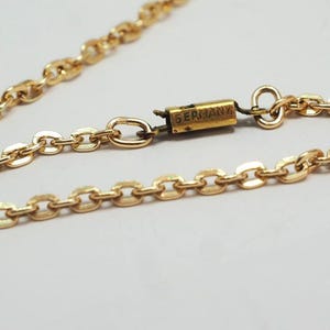 May include: A gold-colored necklace with an oval link chain. The clasp is rectangular with the word "GERMANY" printed on it. The necklace is on a white background.