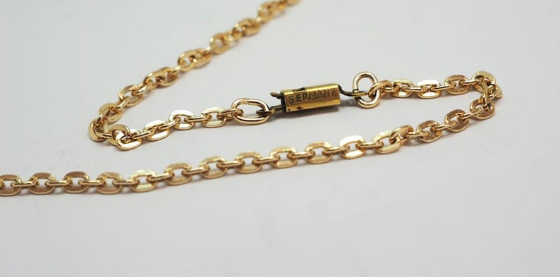 May include: A gold-colored necklace with an oval link chain. The clasp is rectangular with the word "GERMANY" printed on it. The necklace is on a white background.