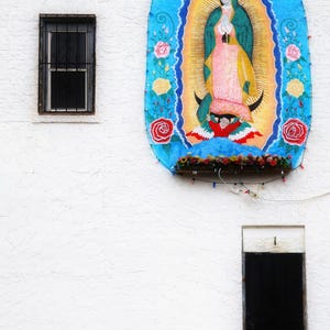 May include: A white building with a mural of the Virgin Mary on the side. The mural is painted in a blue, yellow, red, and green color scheme. The Virgin Mary is wearing a blue robe and a yellow crown. She is holding a baby in her arms. The mural is surrounded by a border of flowers.