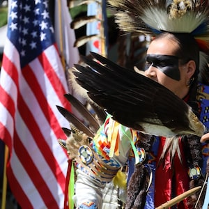 May include: A person in traditional Native American regalia, holding a large feather fan. The outfit includes a feathered headdress, beaded armbands, and colorful ribbons. An American flag is visible in the background.