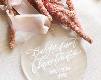 Puede incluir: Adorno redondo de acrílico transparente con el texto "Baby's first Christmas" en letras blancas de escritura. Debajo del texto, "NAVY LOU 2019" en letras blancas en bloque.