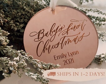 Puede incluir: Un adorno redondo de color rosa con el texto "Baby's first Christmas" en letra cursiva. El nombre "Emily Lynn" y el año "2021" también están escritos en el adorno.