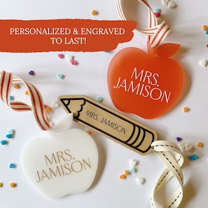 May include: Two personalized apple-shaped teacher name tags, one white and one orange, with the name "MRS. JAMISON" engraved on each. A wooden pencil-shaped tag with the same name is also included. The tags are hanging from white and red striped ribbon and a white ribbon with black stitching.