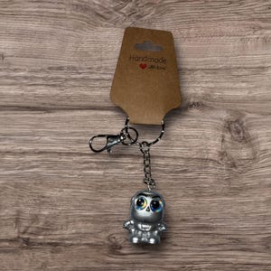 Puede incluir: Un llavero plateado con una peque&ntilde;a figura estilizada con grandes ojos azules. La figura est&aacute; unida a una cadena y a un cierre met&aacute;lico. Una etiqueta marr&oacute;n con las palabras "Handmade with love" est&aacute; unida al llavero.