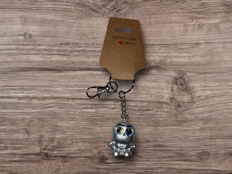 Puede incluir: Un llavero plateado con una peque&ntilde;a figura estilizada con grandes ojos azules. La figura est&aacute; unida a una cadena y a un cierre met&aacute;lico. Una etiqueta marr&oacute;n con las palabras "Handmade with love" est&aacute; unida al llavero.