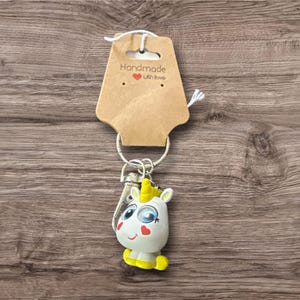 May include: A handmade keychain featuring a white unicorn with blue eyes, a yellow horn, and yellow hooves. The keychain is attached to a metal ring and a brown tag that reads "Handmade with Love."