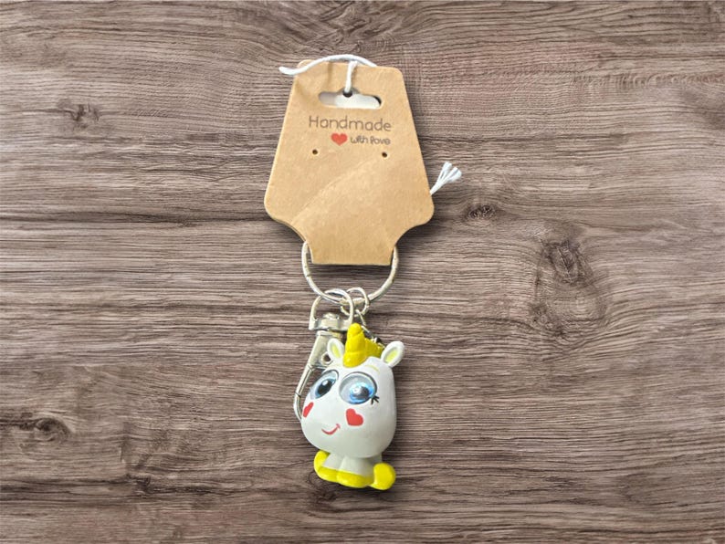 May include: A handmade keychain featuring a white unicorn with blue eyes, a yellow horn, and yellow hooves. The keychain is attached to a metal ring and a brown tag that reads "Handmade with Love."
