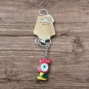 May include: A handmade keychain featuring a red, yellow, and green cartoon character with a large eye. The keychain has a silver chain and clasp, and a brown tag that says "Handmade with love".