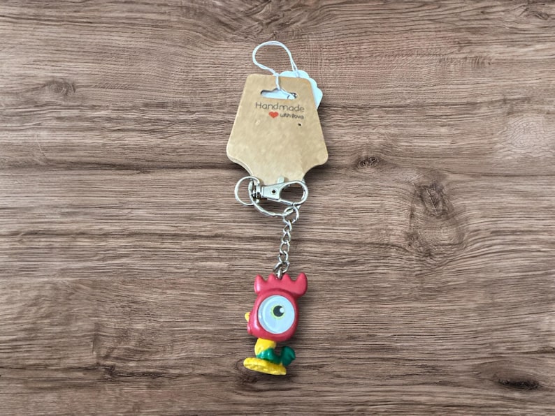 May include: A handmade keychain featuring a red, yellow, and green cartoon character with a large eye. The keychain has a silver chain and clasp, and a brown tag that says "Handmade with love".