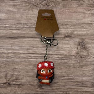 May include: A keychain featuring a small, cartoon-like figure of a girl with dark hair and a red floral headband. The figure is attached to a silver keyring and a brown tag that reads "Handmade with love".