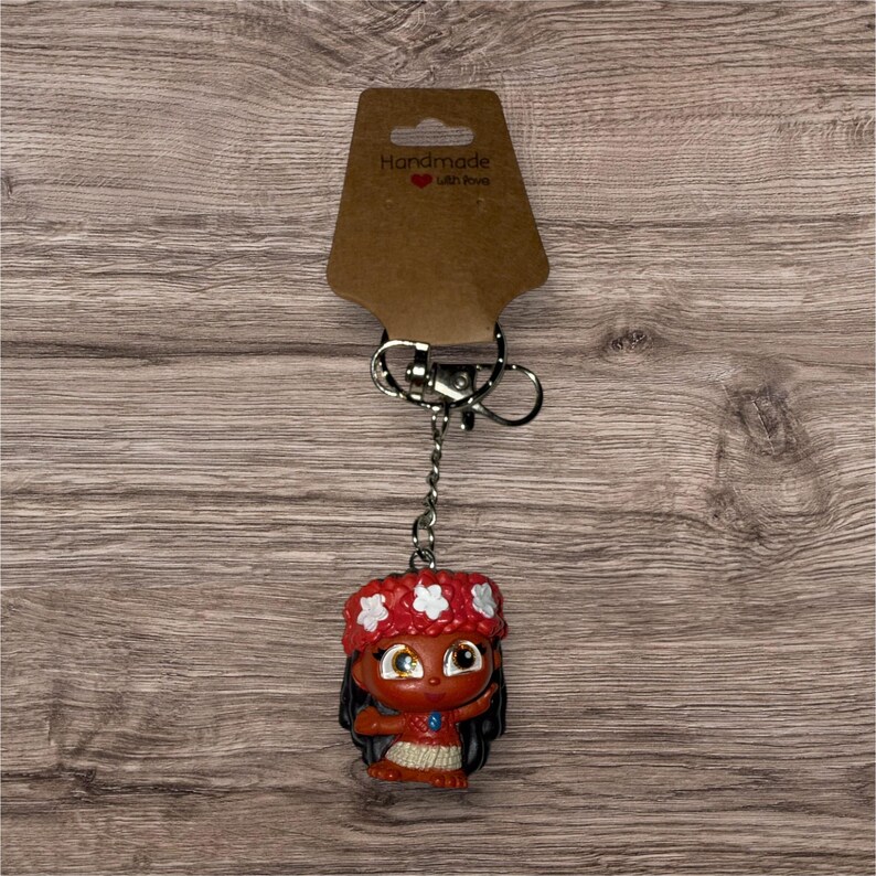 May include: A keychain featuring a small, cartoon-like figure of a girl with dark hair and a red floral headband. The figure is attached to a silver keyring and a brown tag that reads "Handmade with love".