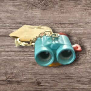 May include: A turquoise-colored miniature binoculars keychain with a gold chain and a small, attached tag. The binoculars have black lenses and a red accent on the side. The keychain is set against a wooden background.