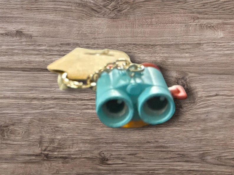 May include: A turquoise-colored miniature binoculars keychain with a gold chain and a small, attached tag. The binoculars have black lenses and a red accent on the side. The keychain is set against a wooden background.
