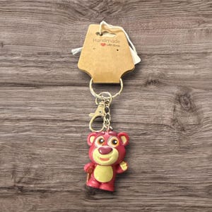 May include: A keychain featuring a pink bear character with a yellow belly and a smiling expression. The keychain is attached to a metal ring and clasp, with a tag that reads "Handmade with love".