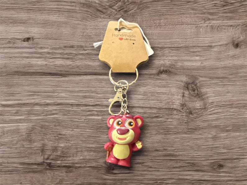 May include: A keychain featuring a pink bear character with a yellow belly and a smiling expression. The keychain is attached to a metal ring and clasp, with a tag that reads "Handmade with love".