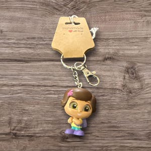 May include: A keychain featuring a cartoon character figurine with a brown bob hairstyle, wearing a yellow top, purple shoes, and a pink bow. The keychain includes a metal clasp and a tag that reads "Handmade with love."