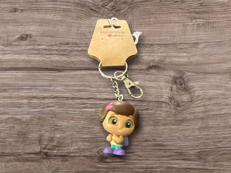 May include: A keychain featuring a cartoon character figurine with a brown bob hairstyle, wearing a yellow top, purple shoes, and a pink bow. The keychain includes a metal clasp and a tag that reads "Handmade with love."