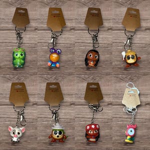May include: A collection of character keychains, each attached to a silver keyring and a brown tag. The keychains feature various colorful cartoon figures, including a green character, a purple-eyed creature, and a character with a red and white outfit.