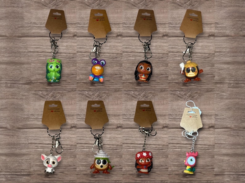 May include: A collection of character keychains, each attached to a silver keyring and a brown tag. The keychains feature various colorful cartoon figures, including a green character, a purple-eyed creature, and a character with a red and white outfit.