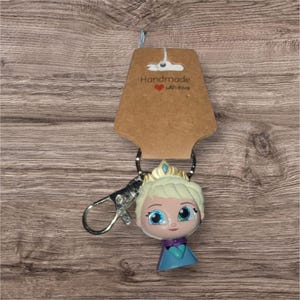 May include: A handmade keychain with a cartoon character wearing a crown and a blue and purple dress. The keychain has a silver clasp and a brown tag that reads "Handmade with love."