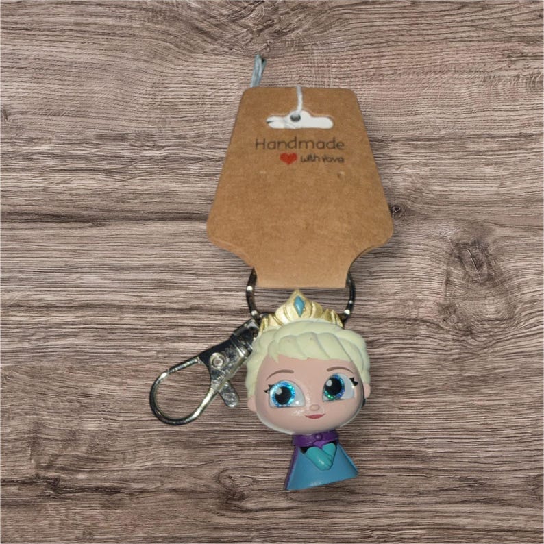 May include: A handmade keychain with a cartoon character wearing a crown and a blue and purple dress. The keychain has a silver clasp and a brown tag that reads "Handmade with love."