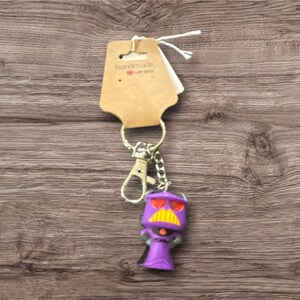 May include: A purple Zurg keychain with a yellow mustache and red eyes. The keychain has a silver clasp and chain, and a brown tag that says "Handmade with love". The background is a wooden surface.