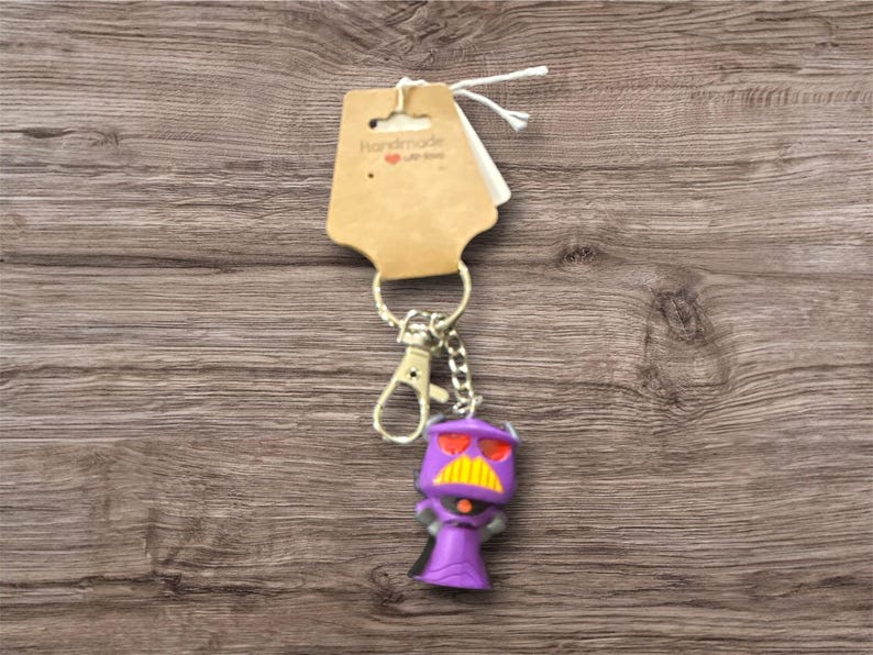 May include: A purple Zurg keychain with a yellow mustache and red eyes. The keychain has a silver clasp and chain, and a brown tag that says "Handmade with love". The background is a wooden surface.