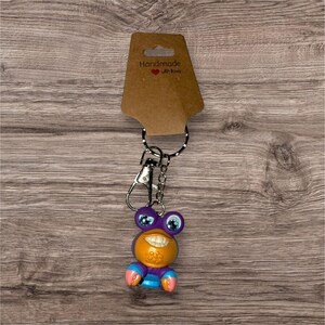 May include: A novelty keychain featuring a smiling, cartoonish creature with purple, orange, blue, and pink accents. The keychain has a silver-colored clasp and a black and white dotted lanyard. The attached tag reads "Handmade with love."