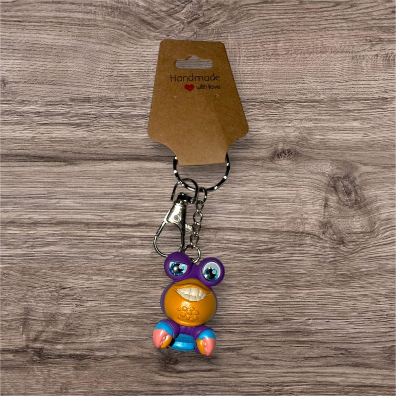 May include: A novelty keychain featuring a smiling, cartoonish creature with purple, orange, blue, and pink accents. The keychain has a silver-colored clasp and a black and white dotted lanyard. The attached tag reads "Handmade with love."