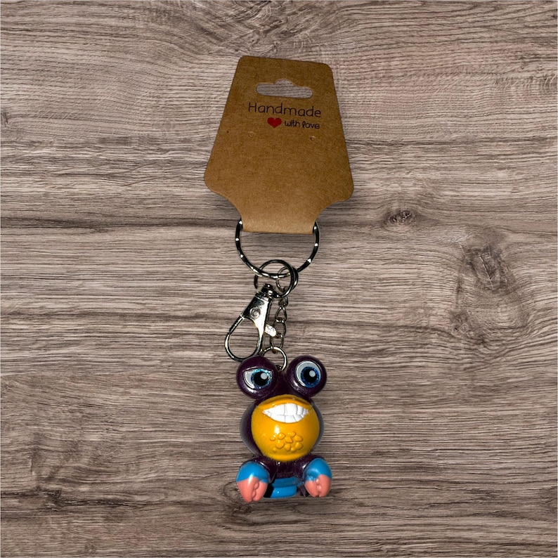 Puede incluir: Un llavero novedoso con un personaje de dibujos animados morado, amarillo y azul con una cara sonriente. El llavero incluye un cierre de metal y un llavero, y est&aacute; unido a una etiqueta marr&oacute;n que dice "Handmade with love."