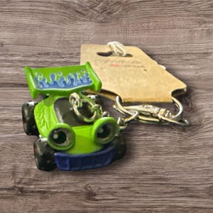 Puede incluir: Un llavero de coche de carreras de juguete verde con un dise&ntilde;o de llamas azules y verdes en el aler&oacute;n. El llavero tiene un cierre plateado y una etiqueta marr&oacute;n adjunta. El coche tiene ruedas negras y detalles azules.