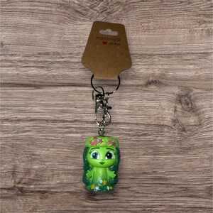May include: A green figurine keychain with a floral crown and a smiling face. The keychain is attached to a silver metal clasp and ring, with a brown tag that reads "Handmade with love". The figurine is approximately 2 inches tall.
