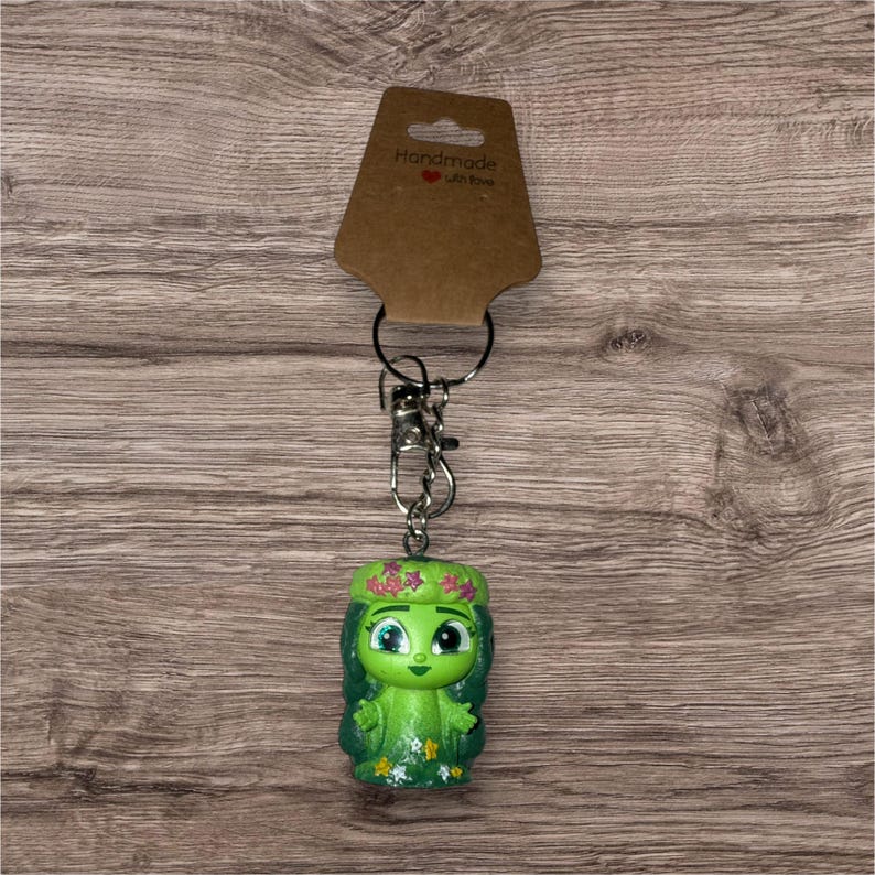 May include: A green figurine keychain with a floral crown and a smiling face. The keychain is attached to a silver metal clasp and ring, with a brown tag that reads "Handmade with love". The figurine is approximately 2 inches tall.