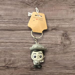May include: A keychain featuring a small figurine of a character wearing a hat and a gray outfit. The keychain includes a metal ring and a brown tag with the text "Handmade with love". The background is a wooden surface.