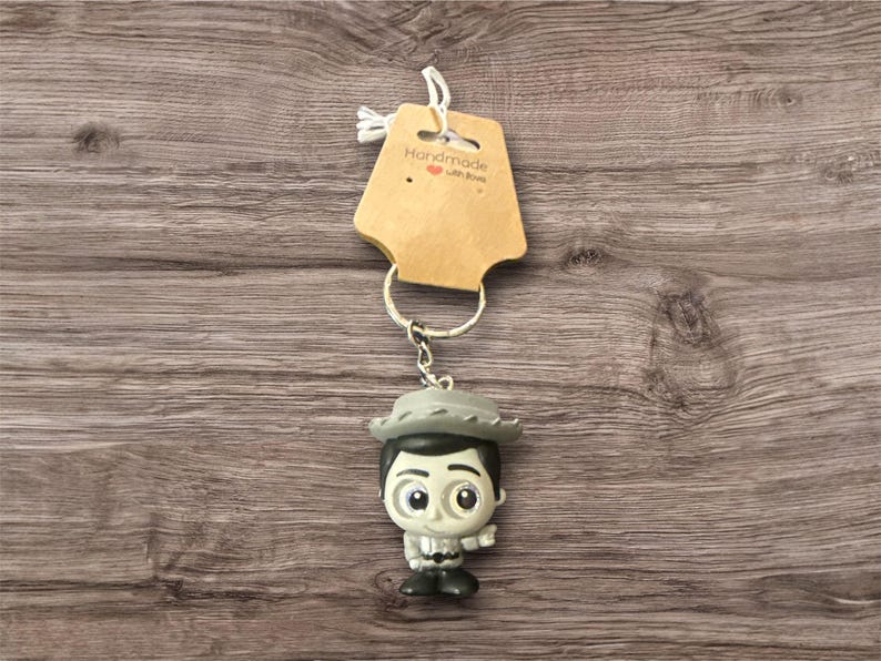 May include: A keychain featuring a small figurine of a character wearing a hat and a gray outfit. The keychain includes a metal ring and a brown tag with the text "Handmade with love". The background is a wooden surface.