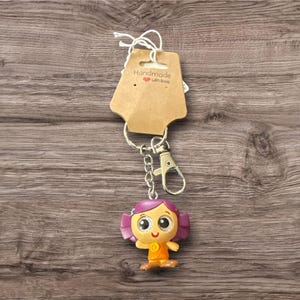 Pu&ograve; includere: Un portachiavi fatto a mano con un personaggio dei cartoni animati con i capelli viola e un vestito arancione. Il portachiavi ha una catena e un fermaglio color argento e un'etichetta marrone con la scritta "Handmade with love".