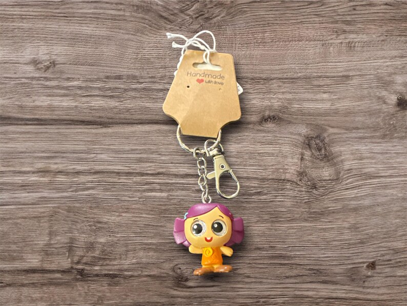 Pu&ograve; includere: Un portachiavi fatto a mano con un personaggio dei cartoni animati con i capelli viola e un vestito arancione. Il portachiavi ha una catena e un fermaglio color argento e un'etichetta marrone con la scritta "Handmade with love".