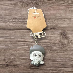 Pu&ograve; includere: Un portachiavi originale con un personaggio con cappello grigio, viso bianco e papillon. Il portachiavi &egrave; attaccato a un fermaglio argentato e a un'etichetta marrone con la scritta "Handmade with Love" con un piccolo cuore rosso.