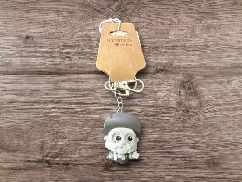 Pu&ograve; includere: Un portachiavi originale con un personaggio con cappello grigio, viso bianco e papillon. Il portachiavi &egrave; attaccato a un fermaglio argentato e a un'etichetta marrone con la scritta "Handmade with Love" con un piccolo cuore rosso.