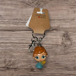 May include: A keyring with a cartoon character figure with orange hair, a blue dress, and a yellow flower. The keyring is attached to a silver clasp and ring, with a brown tag that reads "Handmade with love."