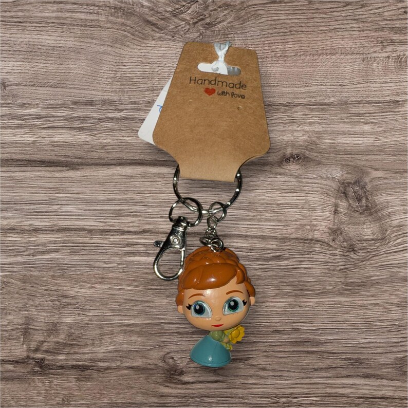 May include: A keyring with a cartoon character figure with orange hair, a blue dress, and a yellow flower. The keyring is attached to a silver clasp and ring, with a brown tag that reads "Handmade with love."