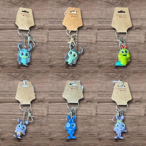 May include: Six handmade keychains featuring colorful cartoon bug figures. Each keychain has a silver-tone ring and clasp, attached to a small, brown tag. The bugs are in various colors, including blue, green, and purple, with detailed features.