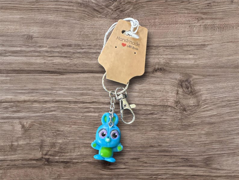 Pu&ograve; includere: Un portachiavi a forma di coniglietto blu e verde con moschettone e portachiavi argentati. Il portachiavi ha un piccolo cartellino marrone con la scritta "Handmade with love". Lo sfondo &egrave; una superficie di legno.
