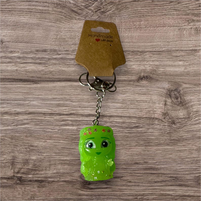 Puede incluir: Un llavero artesanal de resina verde con un personaje de dibujos animados con una corona floral y ojos grandes. El llavero incluye una cadena plateada, un cierre y una etiqueta marr&oacute;n que dice "Handmade with love."
