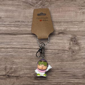 Puede incluir: Un llavero novedoso con un personaje de dibujos animados con gafas de sol verdes, un atuendo blanco y una bufanda rosa. El llavero incluye un cierre y un anillo plateados, sujetos a una etiqueta marr&oacute;n con el texto "Handmade with love."