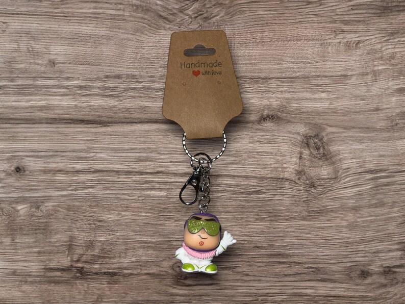Puede incluir: Un llavero novedoso con un personaje de dibujos animados con gafas de sol verdes, un atuendo blanco y una bufanda rosa. El llavero incluye un cierre y un anillo plateados, sujetos a una etiqueta marr&oacute;n con el texto "Handmade with love."