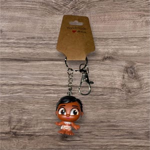 Puede incluir: Un llavero con una peque&ntilde;a figura de un personaje de dibujos animados con pelo oscuro y piel morena. La figura est&aacute; unida a una cadena y un cierre plateados, con una etiqueta marr&oacute;n que dice "Handmade with love".