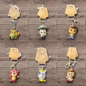 May include: Collection of character keychains with brown tags. Keychains feature various cartoon characters in vibrant colors, including a bear, a unicorn, and a lion. Each keychain is attached to a silver metal clasp. The background is a wooden surface.