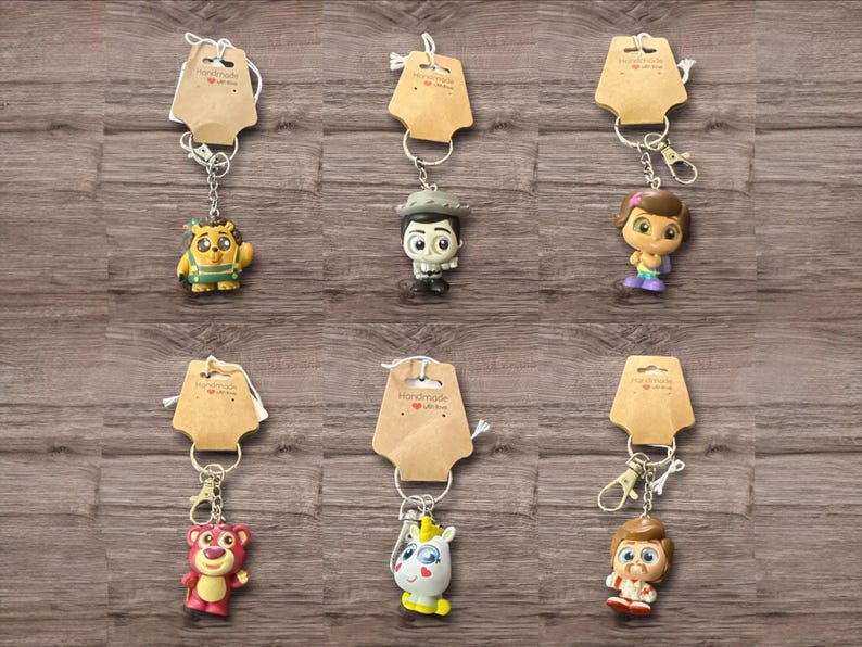 May include: Collection of character keychains with brown tags. Keychains feature various cartoon characters in vibrant colors, including a bear, a unicorn, and a lion. Each keychain is attached to a silver metal clasp. The background is a wooden surface.