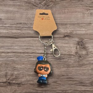 Puede incluir: Un llavero con una peque&ntilde;a figura de una persona con gafas, un sombrero azul y un atuendo azul. El llavero incluye un cierre y un anillo plateados, sujetos a una etiqueta marr&oacute;n que dice "Handmade with love."